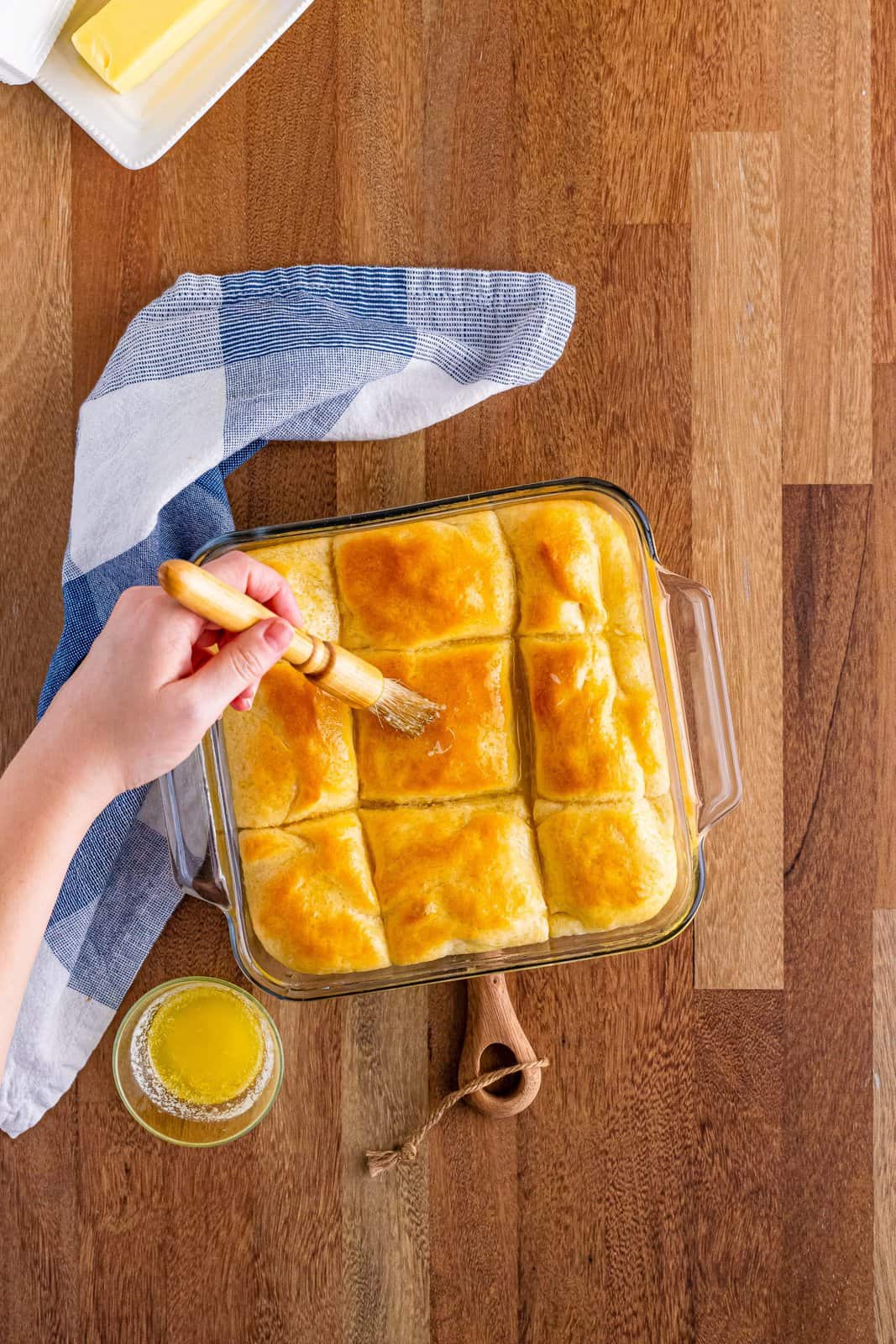A baking dish with fresh, hot butter dip yeast rolls with a brush brushing more butter.