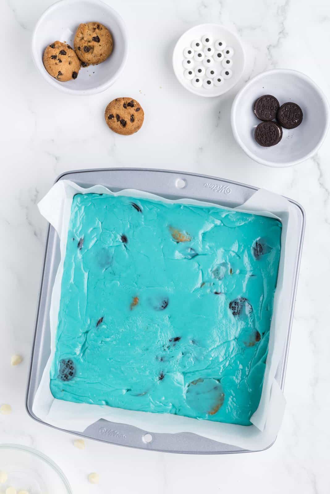 The blue layer of fudge with some cookies mixed in in a baking dish and cookies and candy eyes in other containers around it.