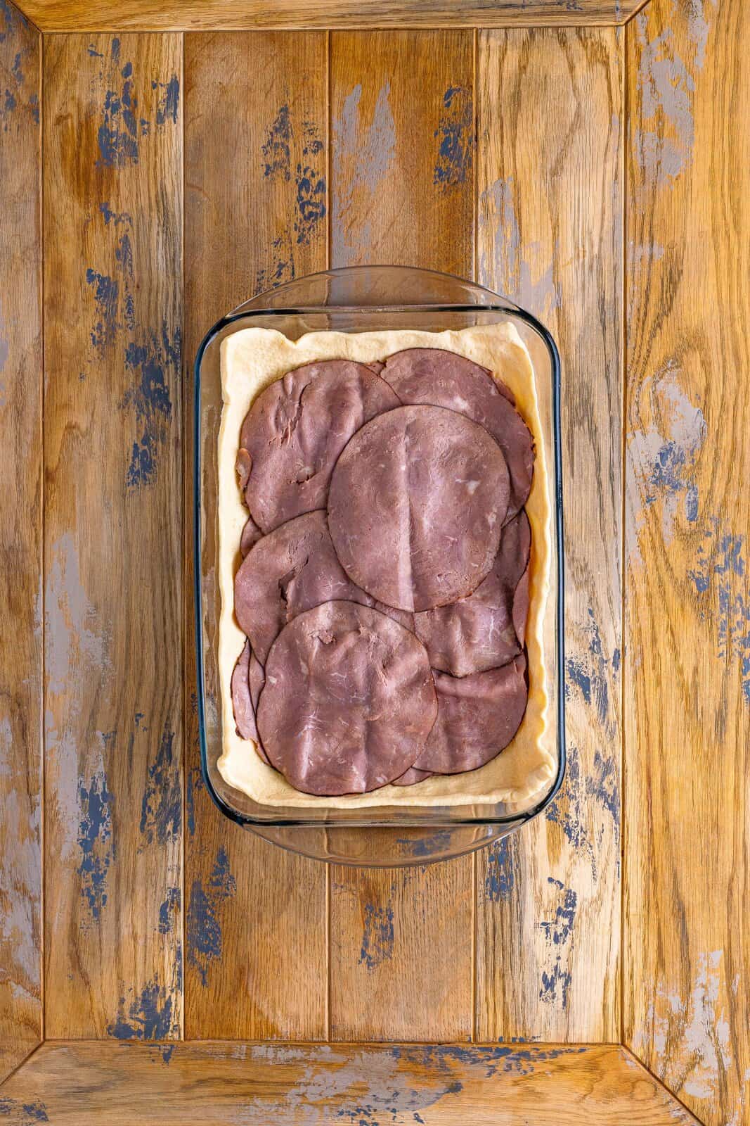 A baking dish with dough, cheese, and roast beef. 