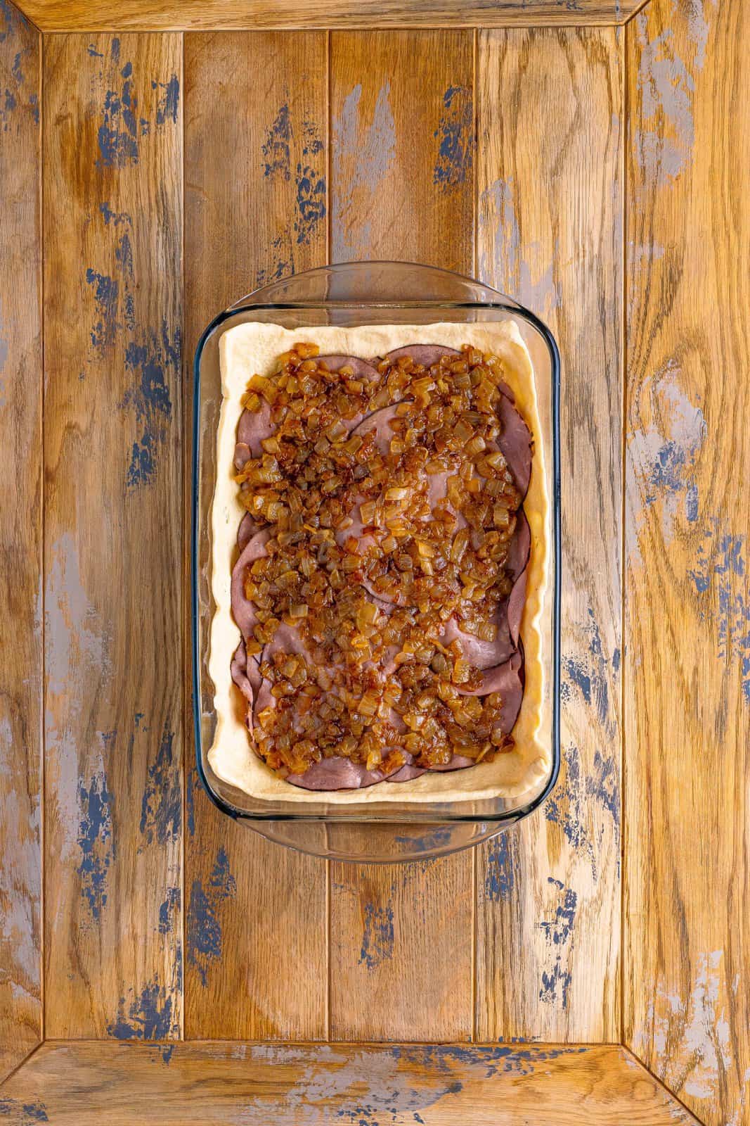 A baking dish with dough, cheese, roast beef, and cooked onions. 