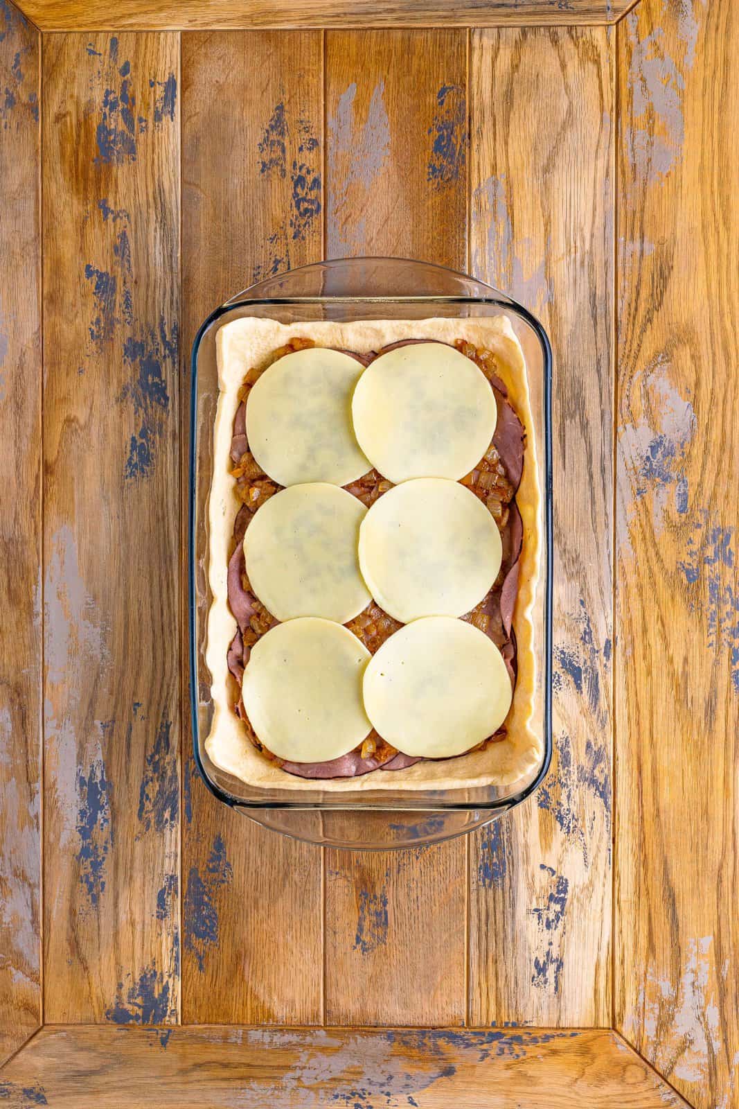 A baking dish layered with dough, cheese, roast beef, cooked onion, and provolone cheese. 