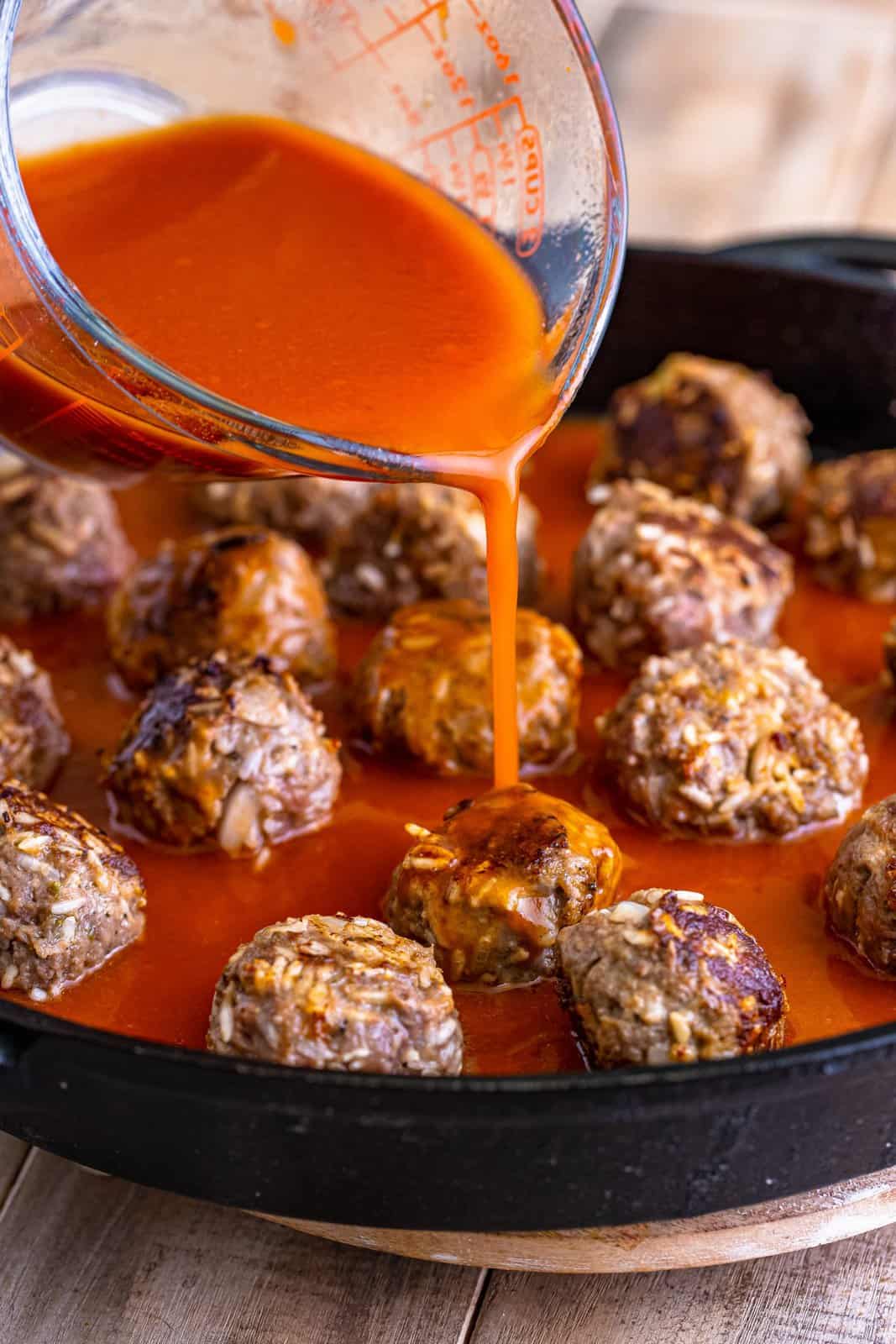 A glass container pouring a tomato sauce over top of the Porcupine Meatballs in a cast iron skillet.