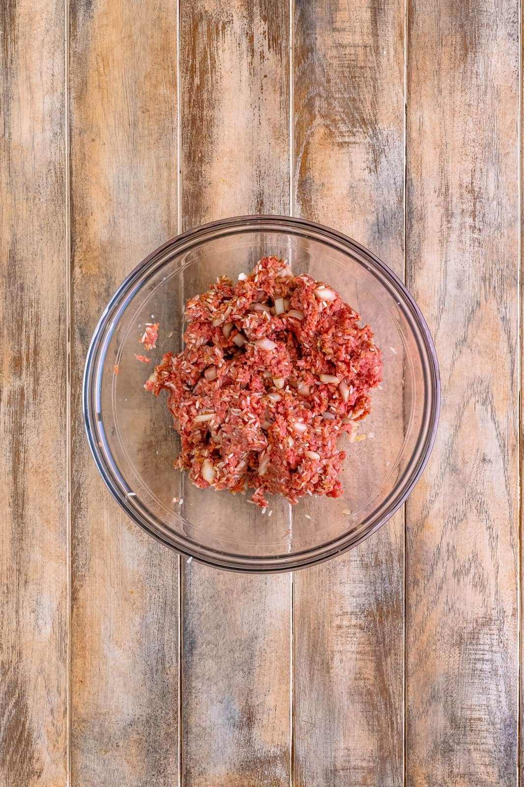 A glass mixing bowl of ground beef and other ingredients needed to make meatballs all mixed together.