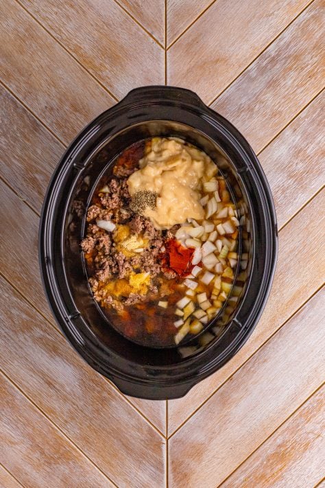 A crockpot insert full of macaroni pasta, herbs and spices, cooked ground beef, beef stock, onion, and can of potato soup.