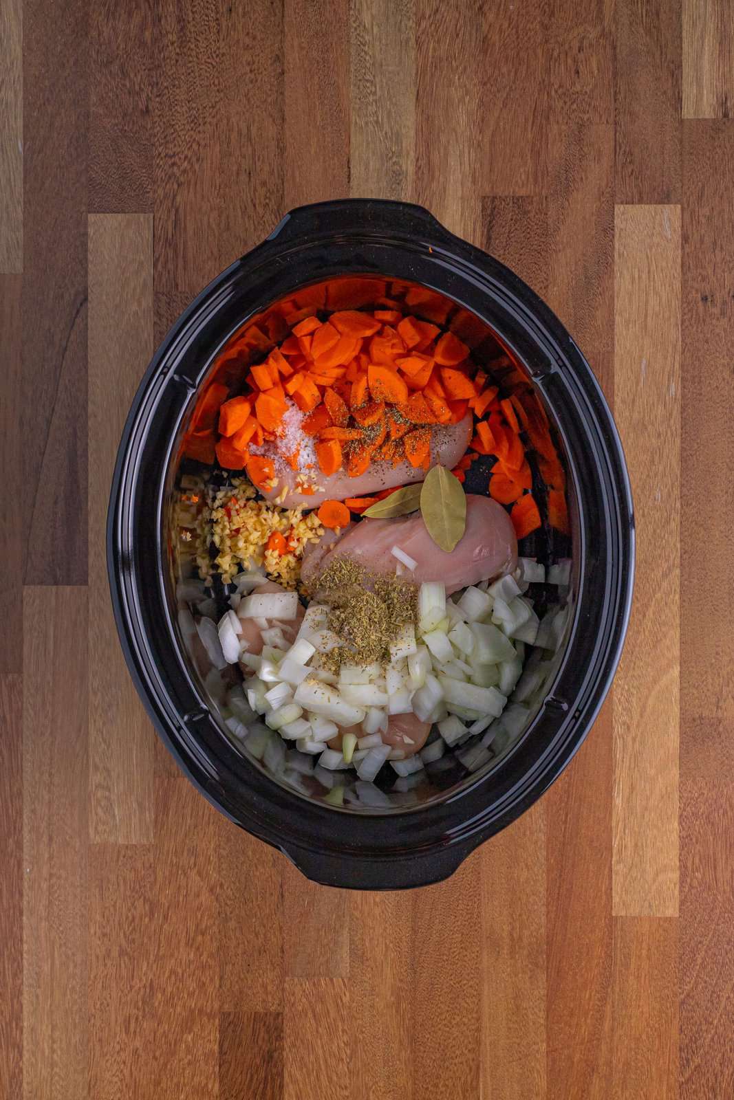 Looking down on a Slow Cooker insert with chicken breasts, carrots, onion, garlic, bay leaves, Italian seasoning, salt, and pepper.