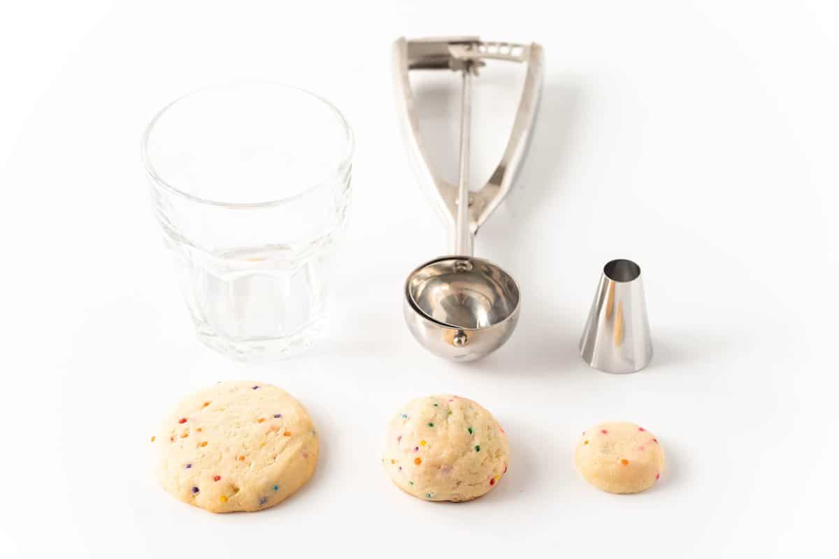 A glass, cookie scooper, and piping tip with a few cookies sitting in front.