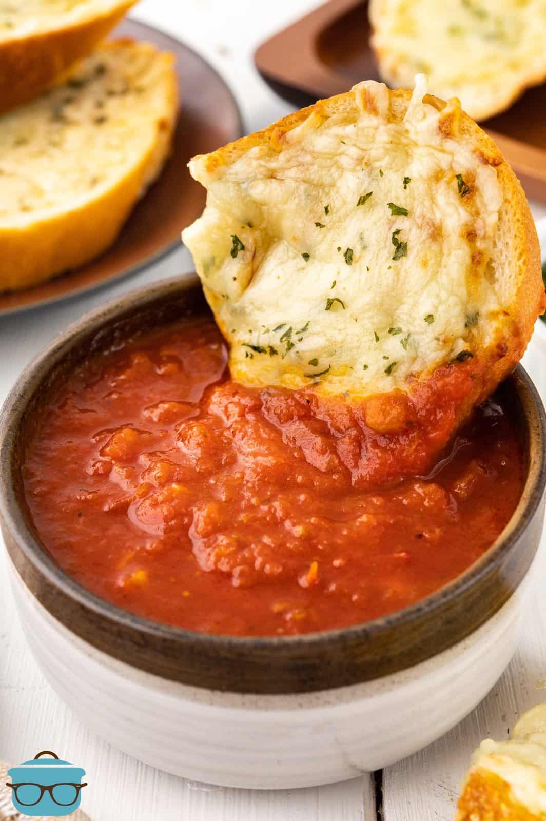 A piece of garlic bread being dipped in a bowl of marinara sauce.