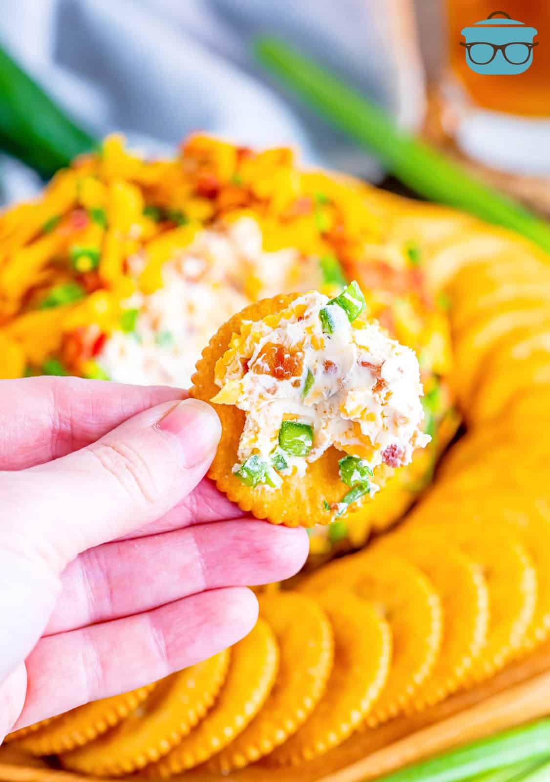 A hand holding a cracker with a serving of Jalapeno cheeseball on it.
