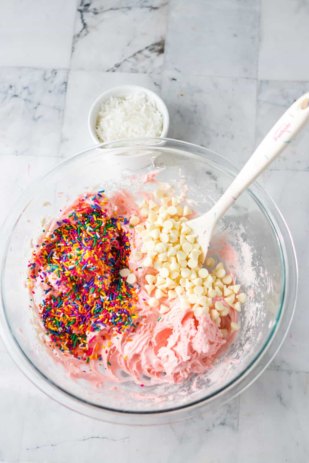 A mixing bowl with cream cheese, strawberry cake mix, sprinkles, and white chocolate chips.