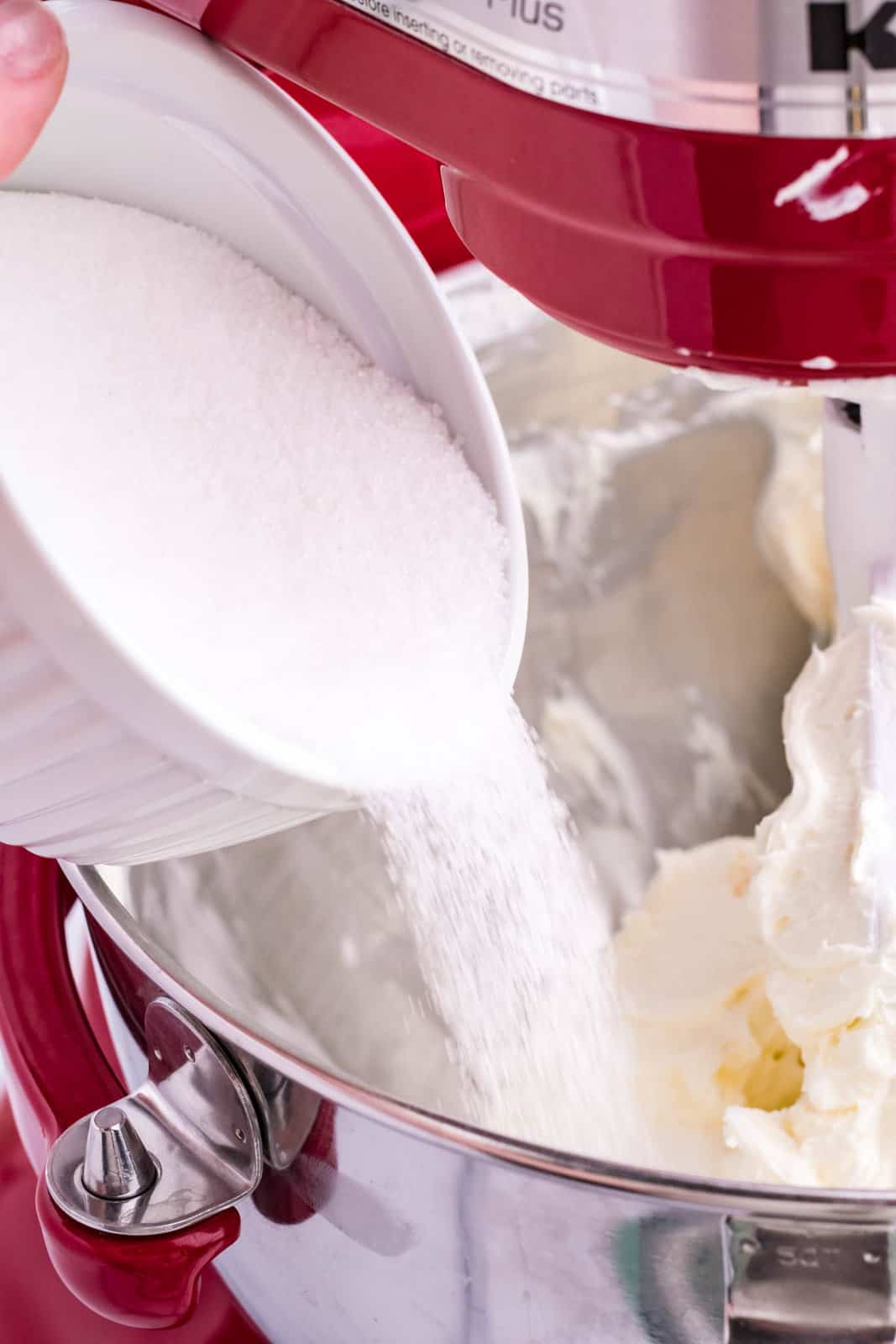 A cup pouring in sugar into a mixing bowl with creamed cream cheese.