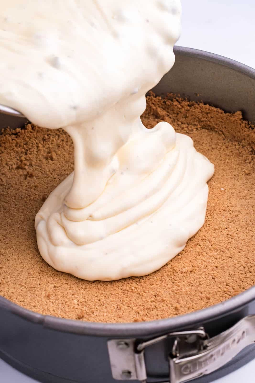 Cheesecake filling being poured into a graham cracker crumb crust in a pan.