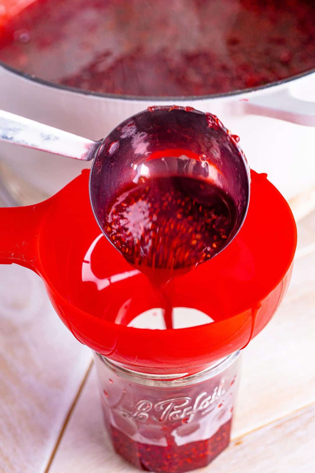 Ladle of raspberry jam being poured through a wide mouth funnel into a jam jar.