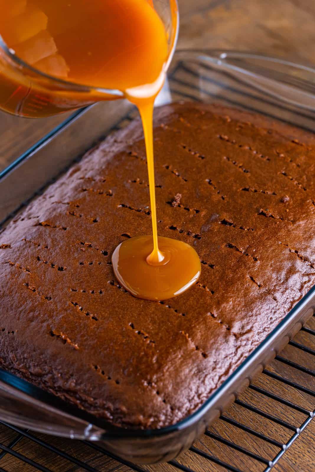 Caramel topping being poured on a chocolate cake with fork holes.
