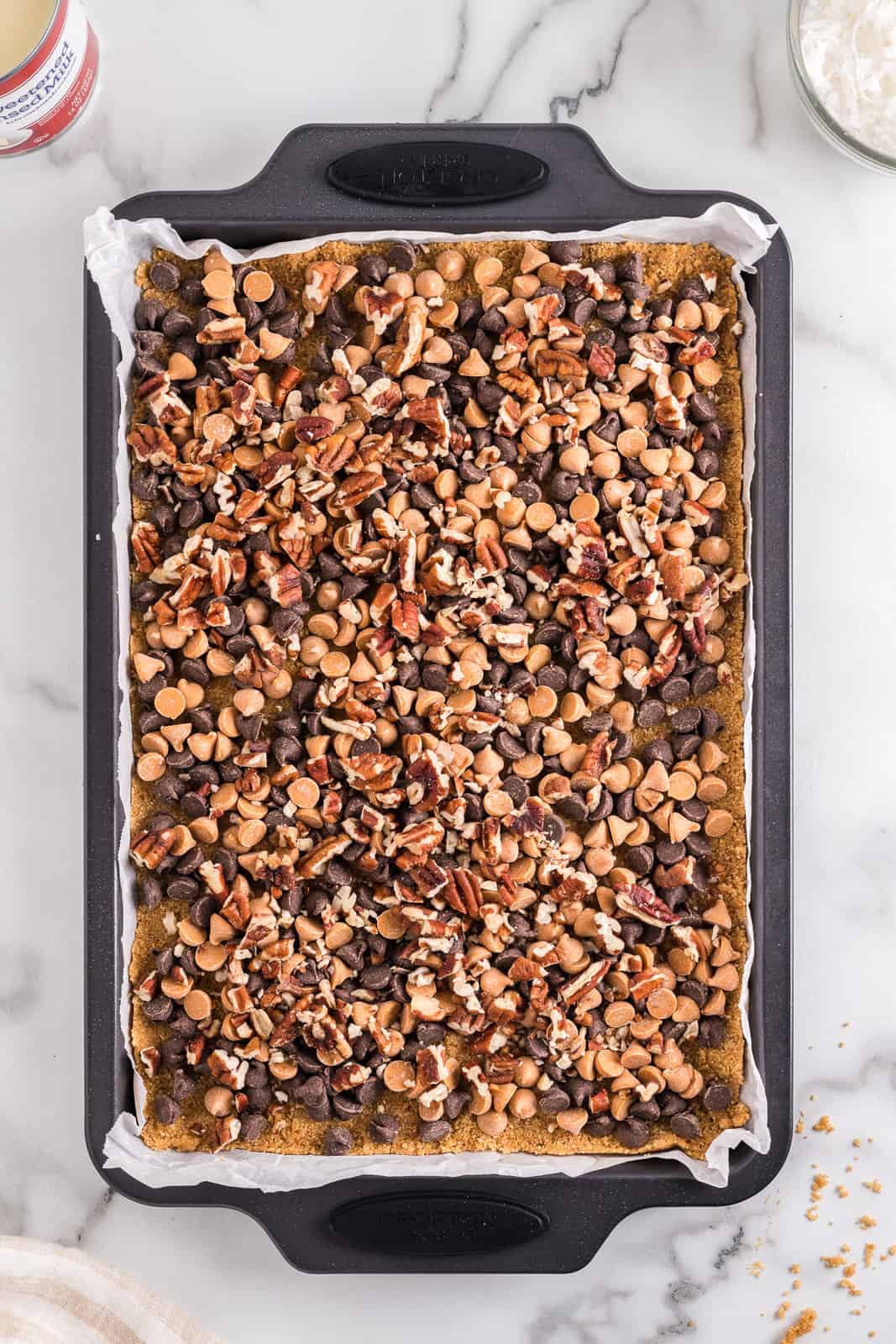 Butterscotch chips, chocolate chips, and pecans on top of a crust on a baking sheet lined with parchment paper.