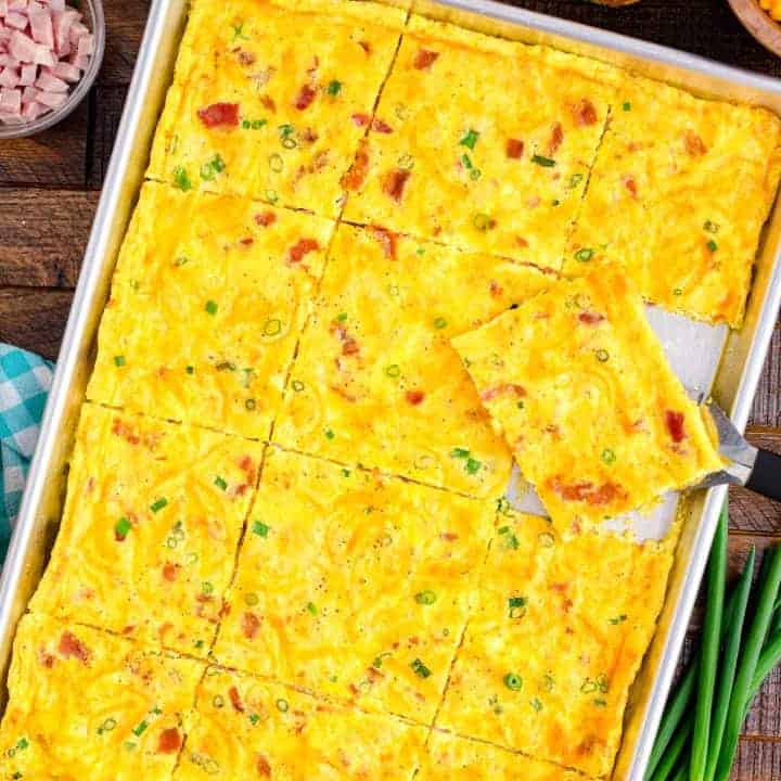 A baking tray with cooked eggs with bacon, ham, and green onions and cheese with a piece cut out.