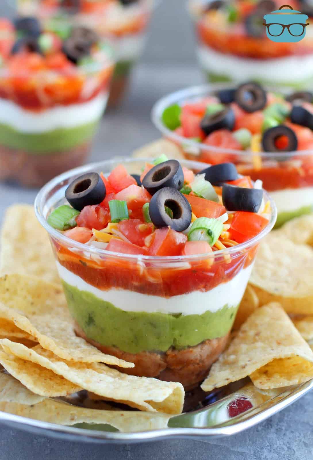 A plastic cup with a layered dip and chips around it on a plate. 