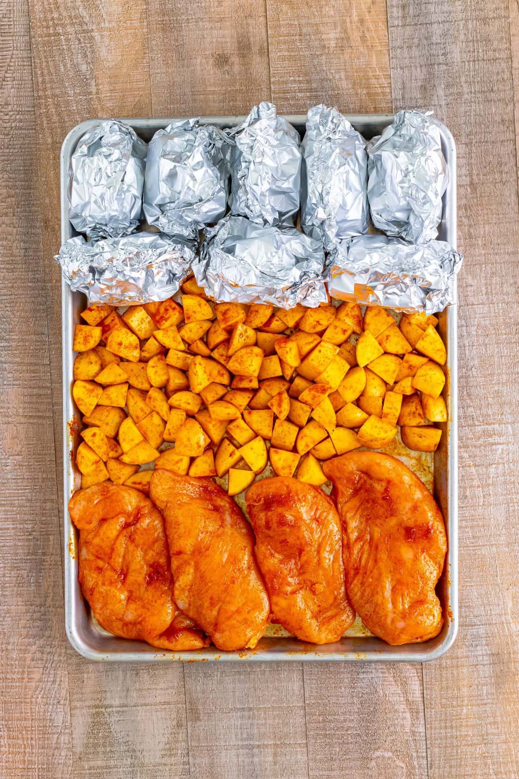 A sheet pan with seasoned chicken, diced potatoes, and aluminum foil covered corn on the cob.