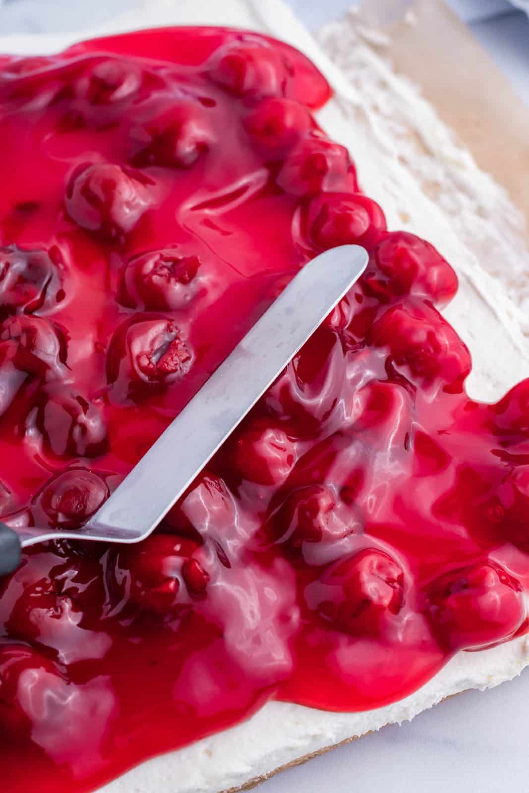 A cherry pie filling being spread out over a whipped cream mixture in a baking dish.