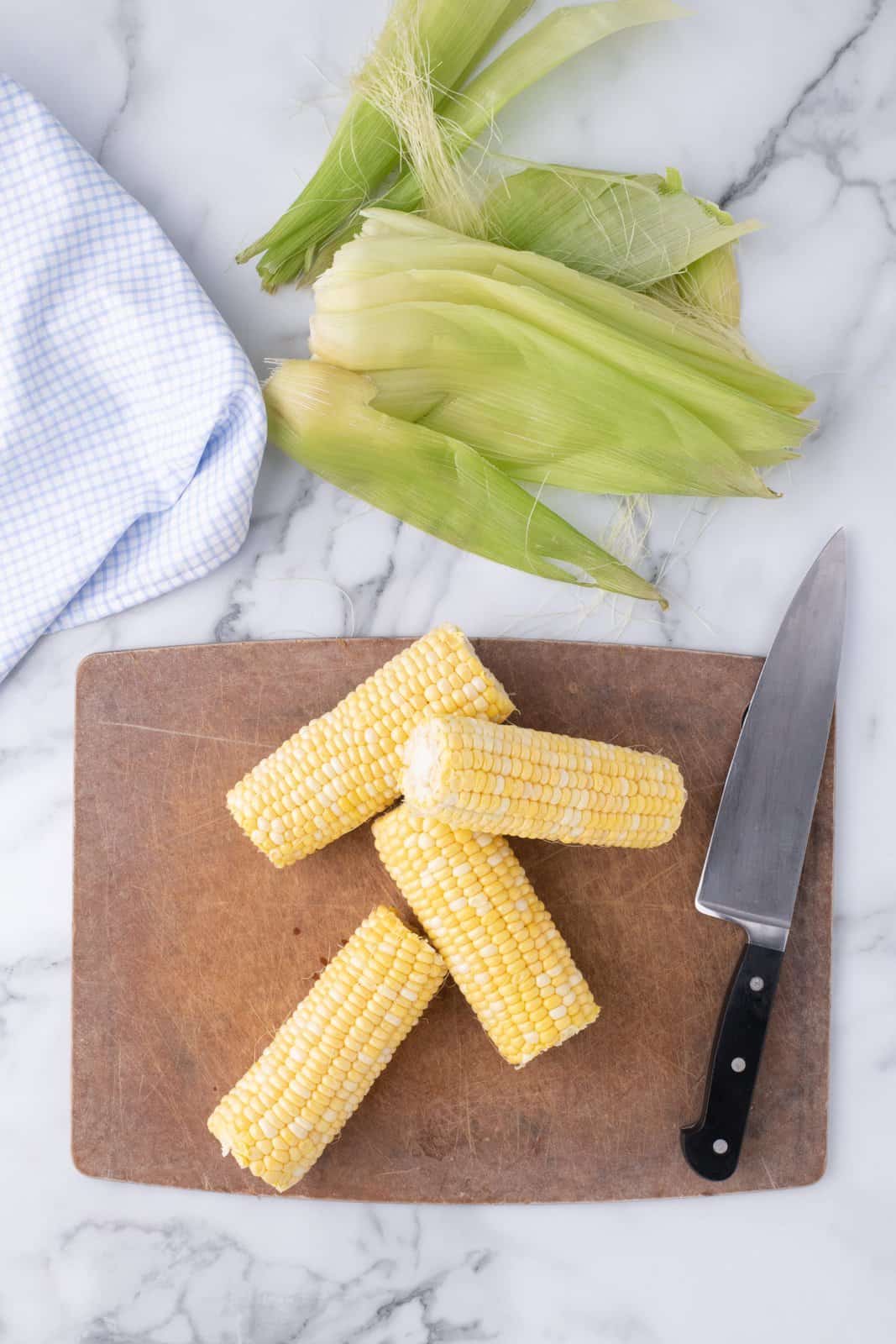 Trimmed ears of corn on a cutting board with knife and corn husks.