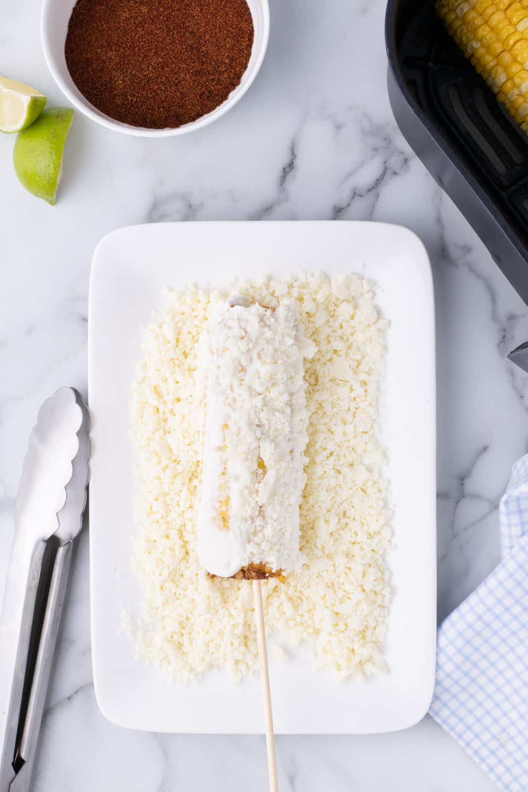 A cob of mayo and sour cream smothered corn being rolled in cotija cheese.