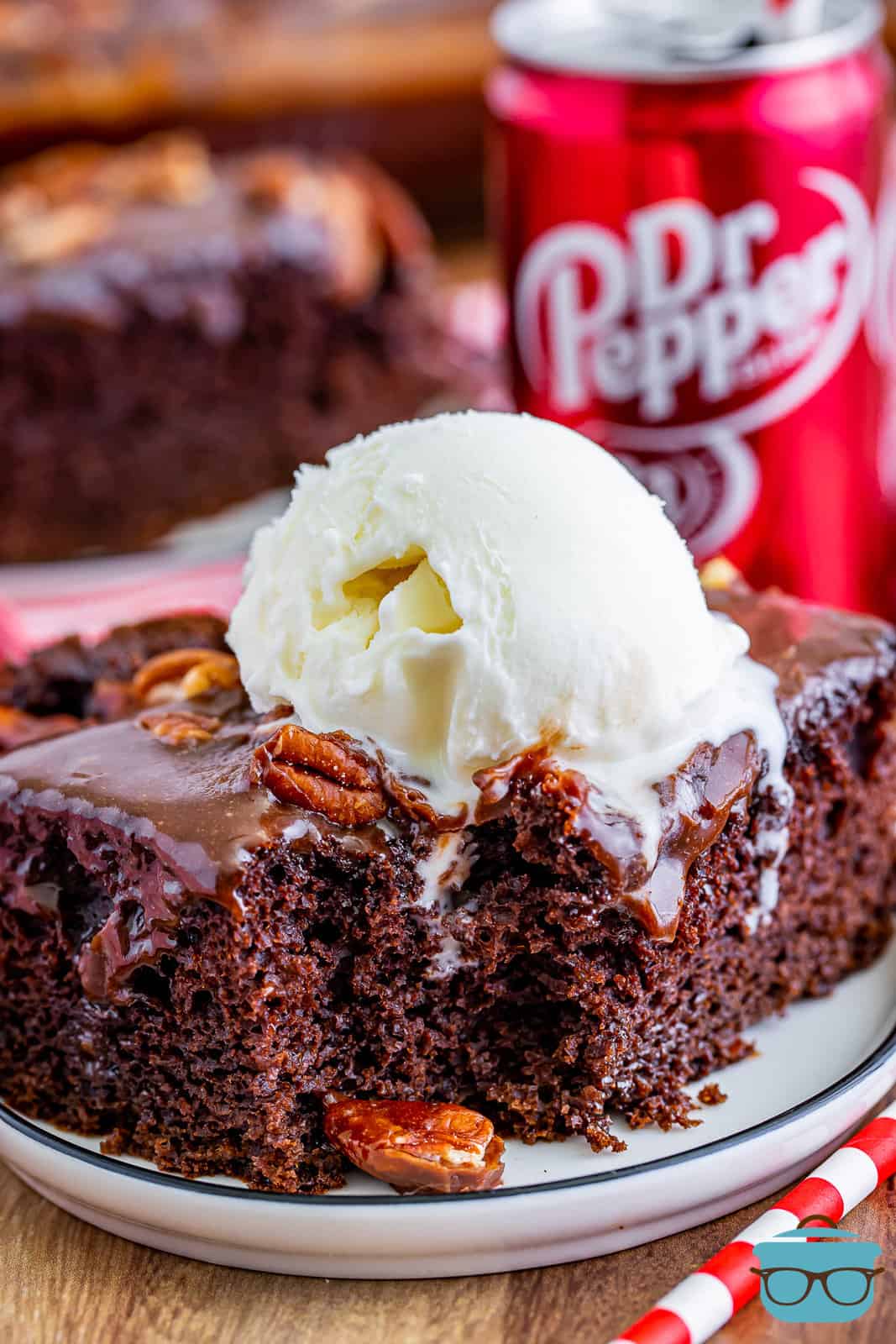 slice of Dr Pepper cake on a small round white plate with a coop of vanilla ice cream slightly melted on top of the cake and a small can of Dr Pepper soda in the background. 