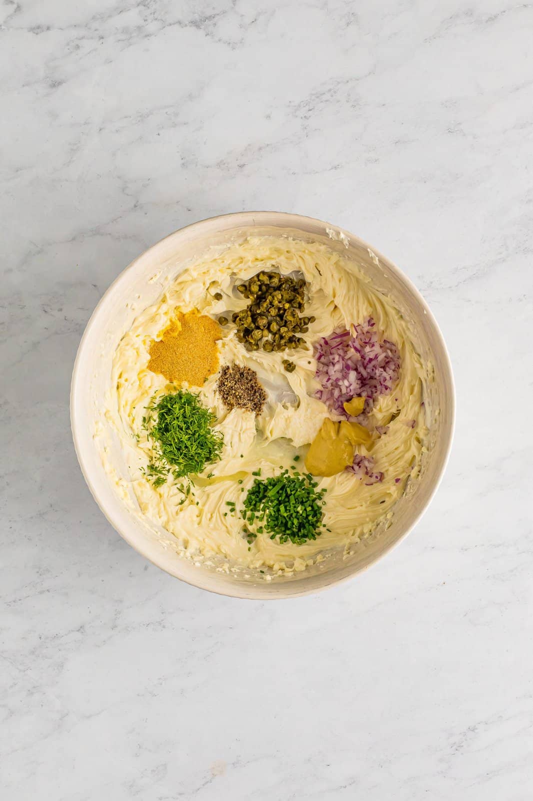 A mixing bowl with cream cheese, sour cream, mayo, diced red onion, dijon mustard, capers, dill, chives, lemon juice, garlic powder, salt and pepper.