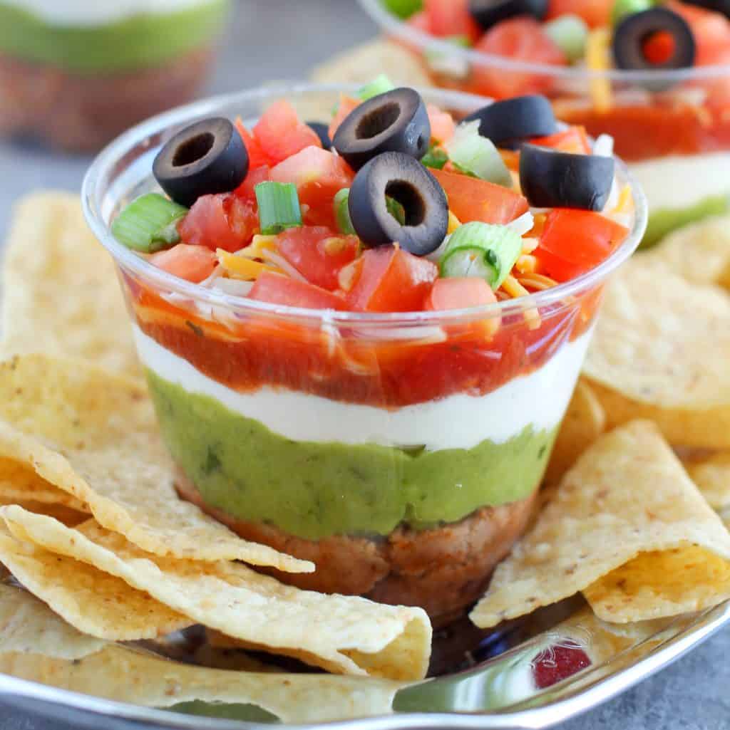 Seven layer dip in clear plastic cups with chips on a cup.