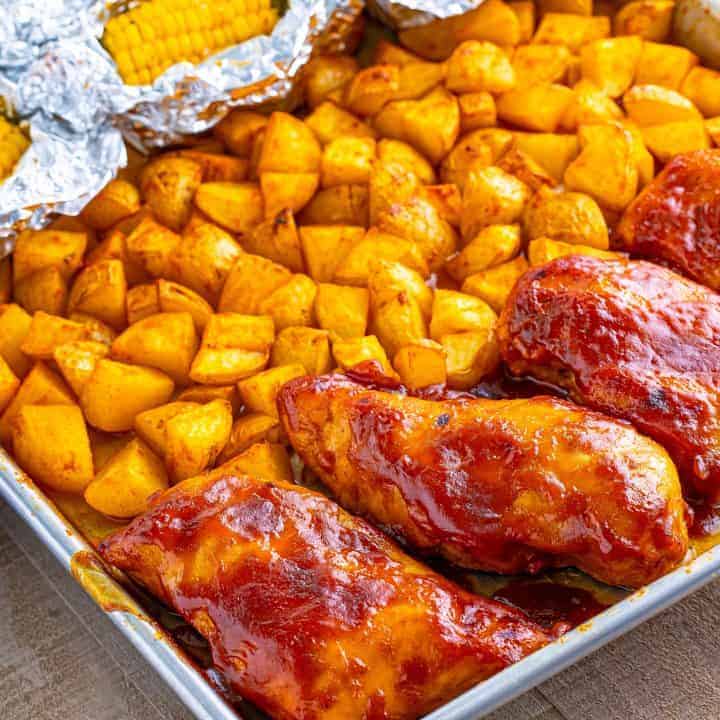 Looking closer at a sheet pan with cooked BBQ chicken, potatoes, and corn.
