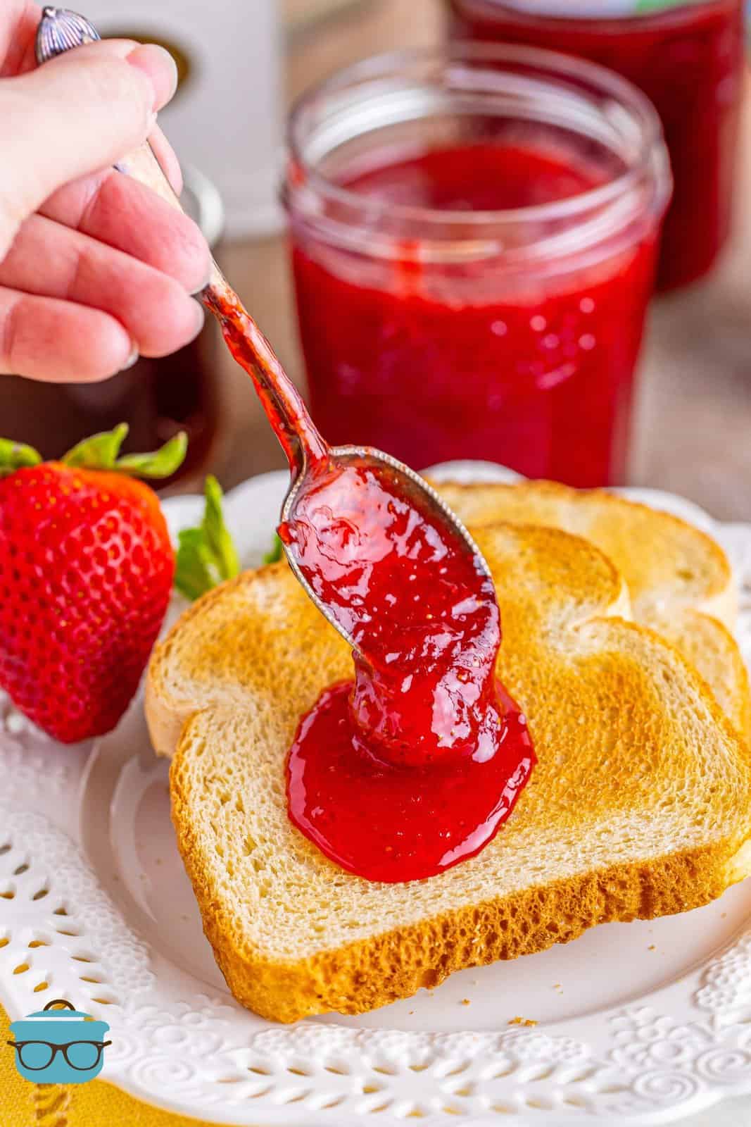 A hand putting a spoonful of strawberry jam on a piece of toast.