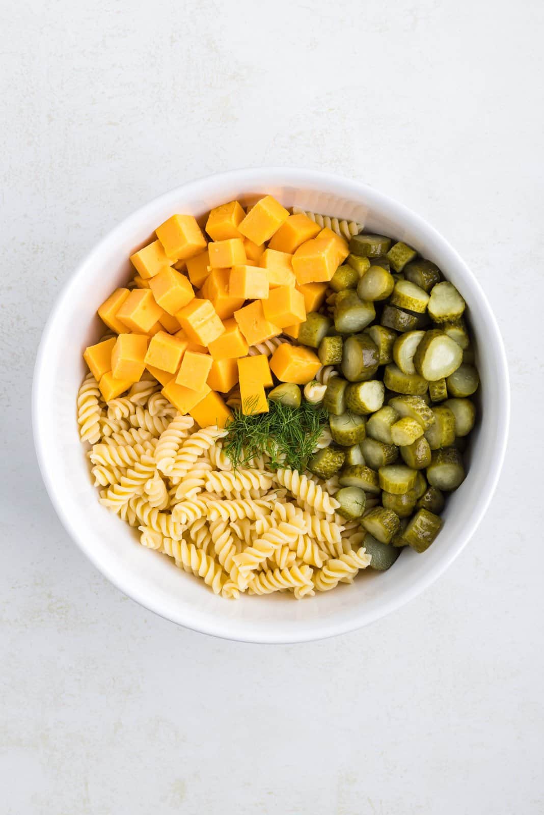 Cooked pasta, cheese, pickles and dill in a mixing bowl. 