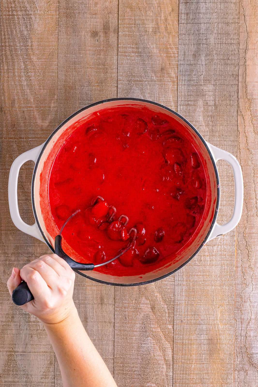A pot of strawberry jam with a potato masher. 
