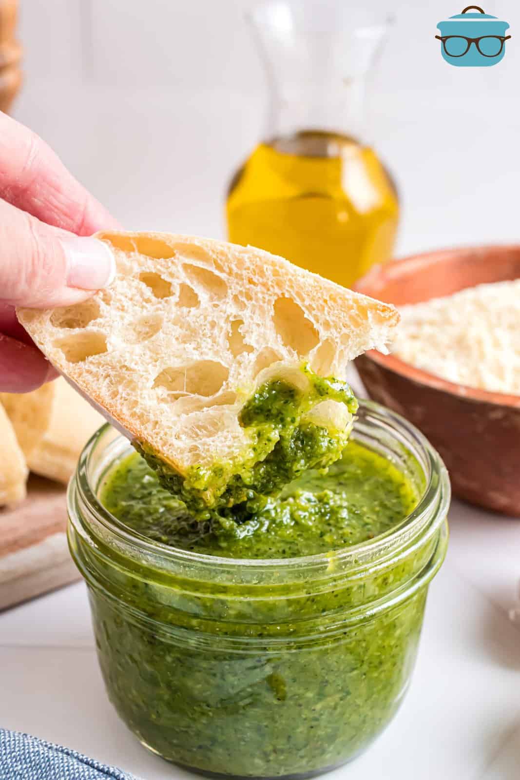 A piece of dipping bread with some basil pesto on it over a jar of it.