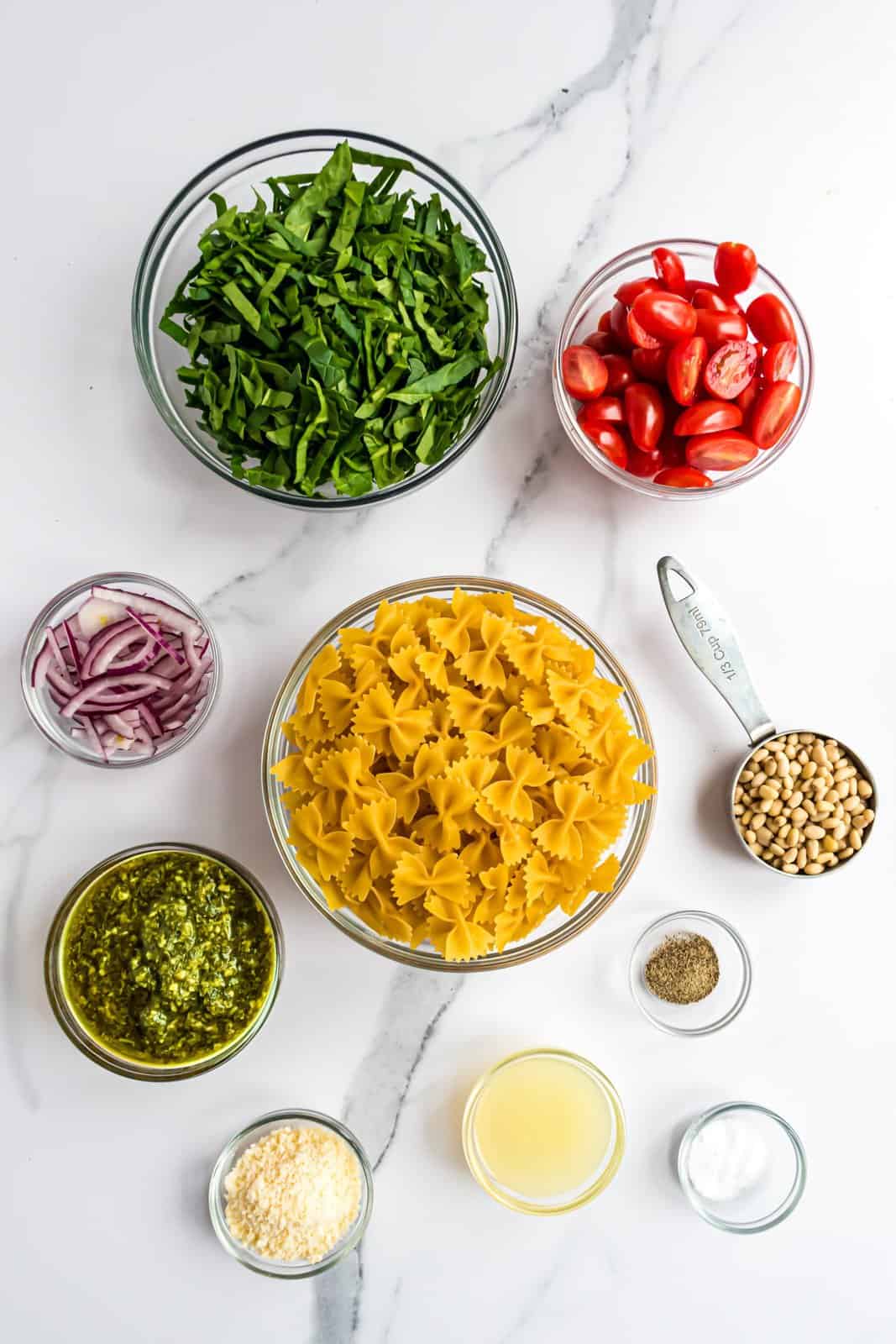 Pine nuts, bow tie pasts, pesto sauce, lemon juice, kosher salt, black pepper, fresh spinach, cherry or grape tomatoes, red onion, and grated or shaved parmesan cheese.