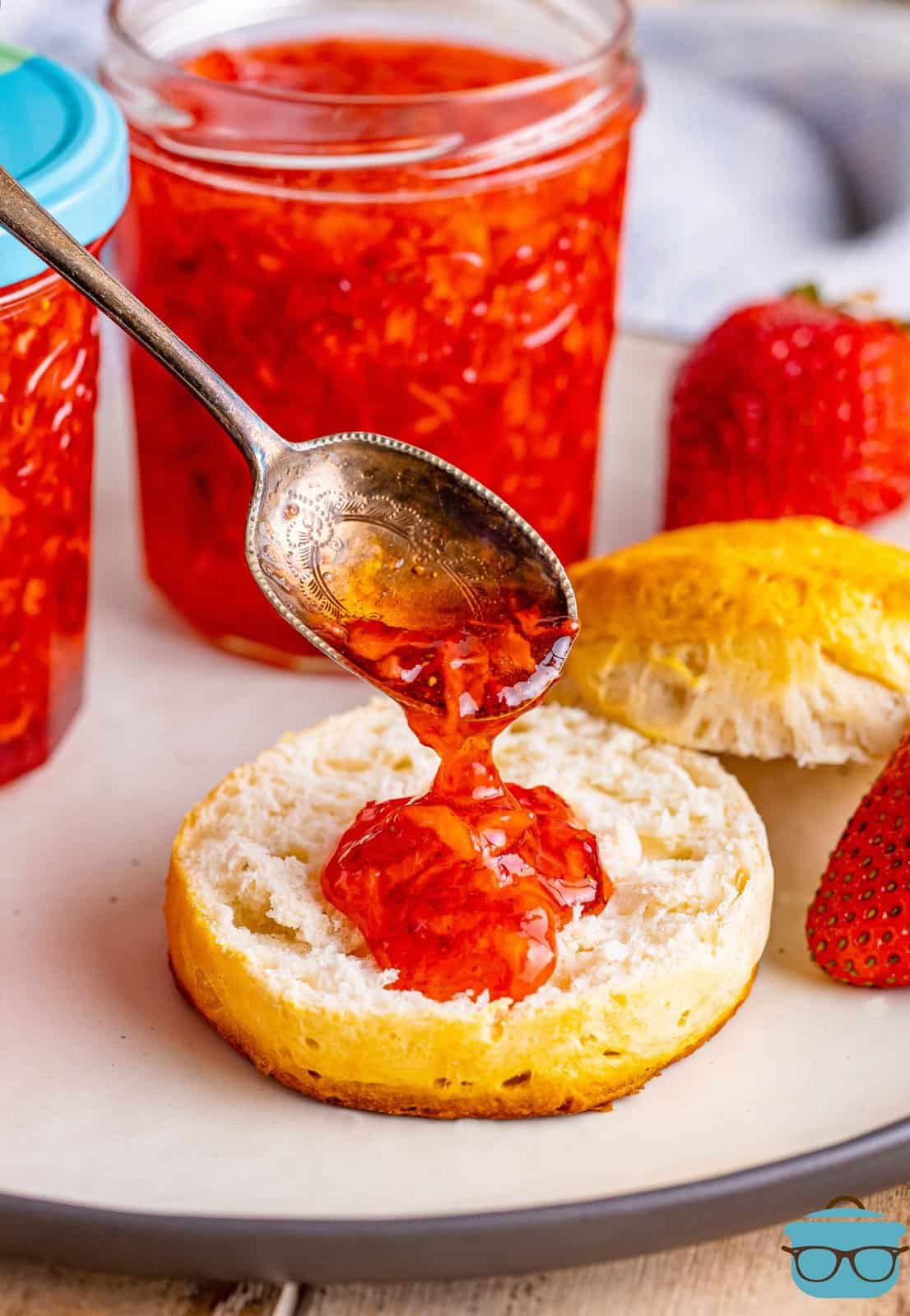 A spoon pouring some strawberry jam on the biscuit.