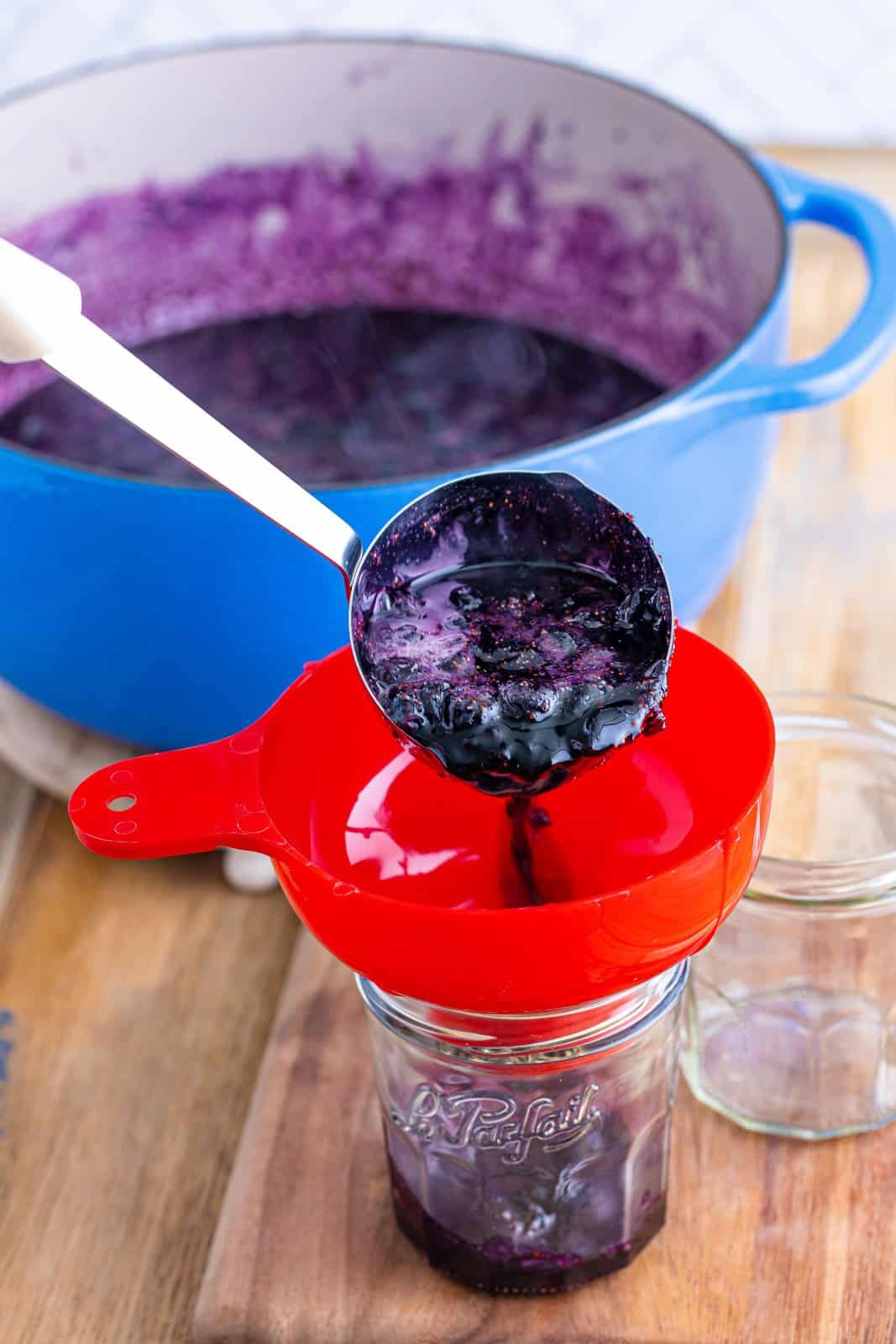 A ladle pouring homemade blueberry jam into a funnel into a jar.
