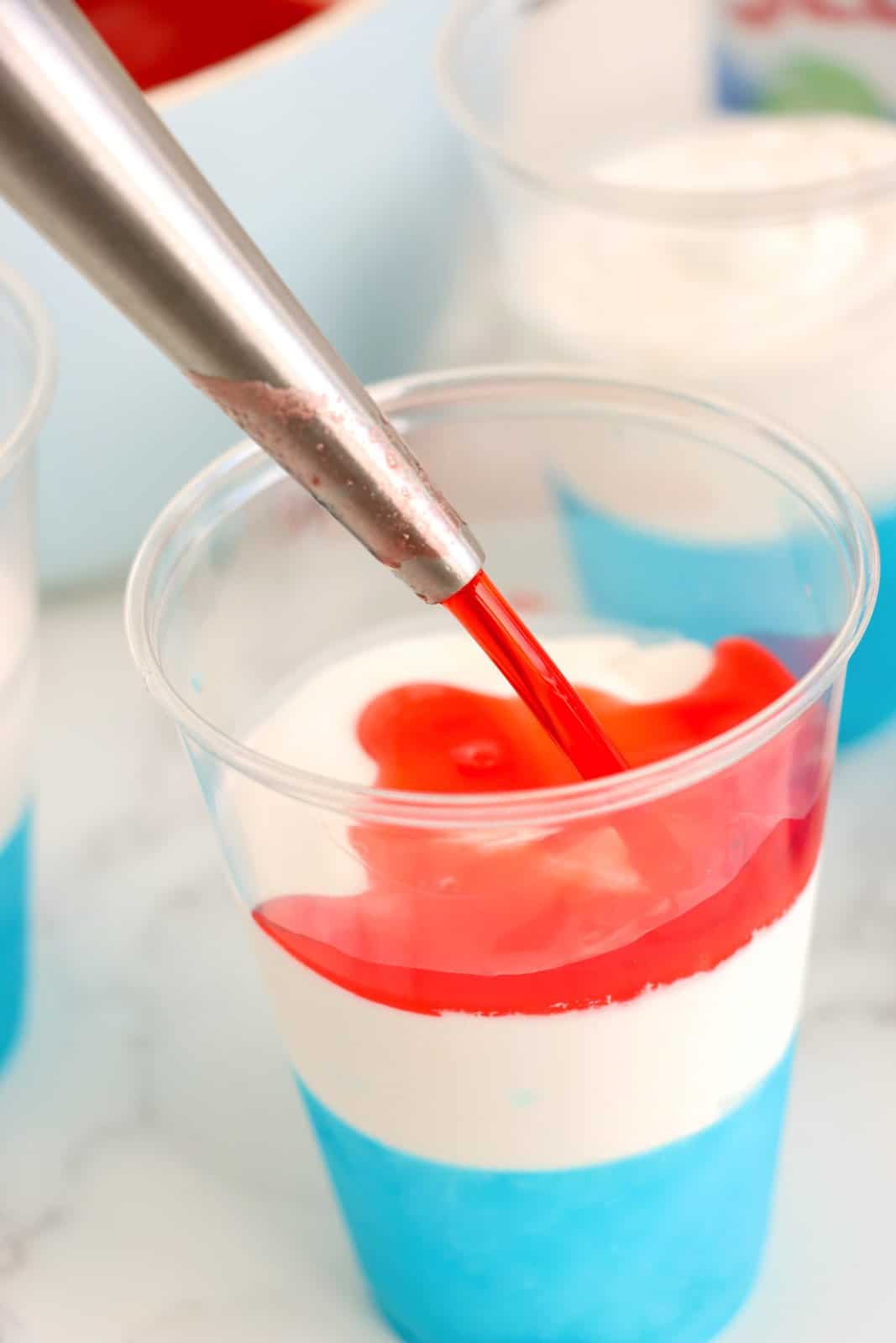 A turkey baster pouring red jello on top of whipped cream and blue jello.