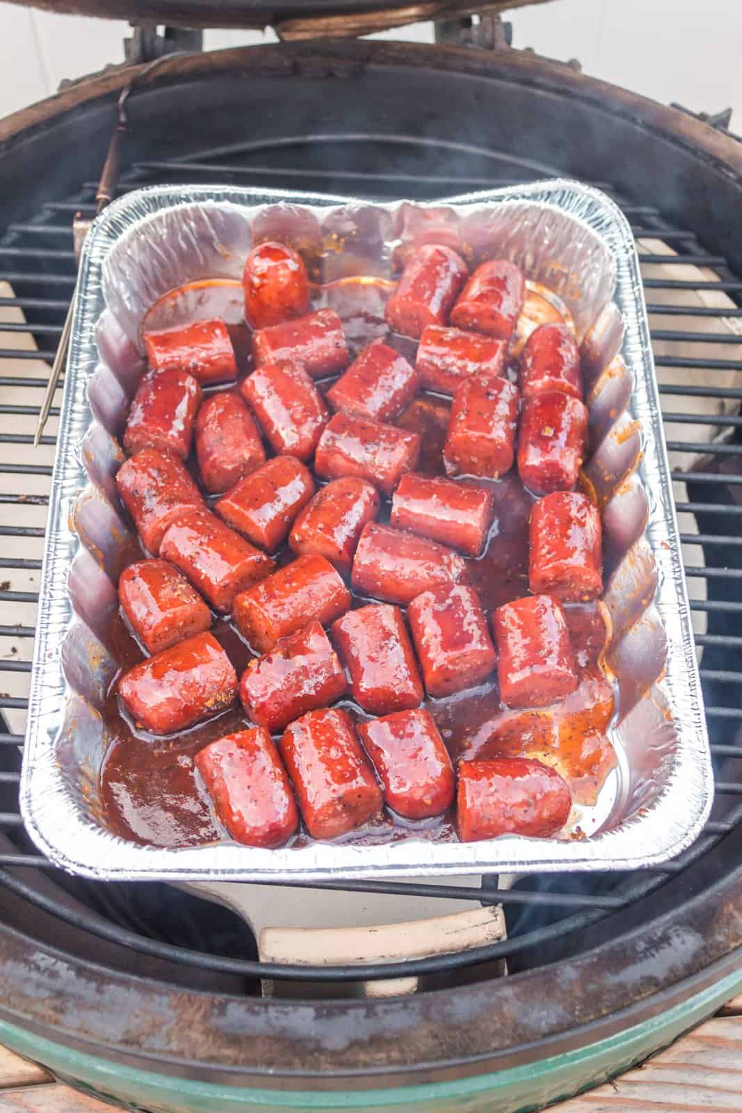 Cut up hot dogs with seasoning and sauce in an aluminum pan on a grill.