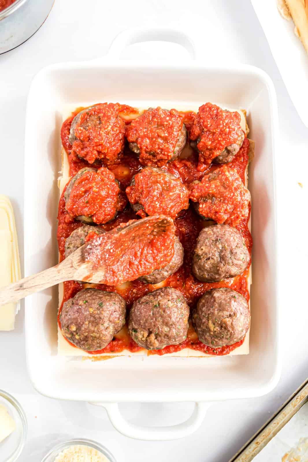 Meatballs on slider rolls with cheese and a wooden spoon spreading sauce on top.