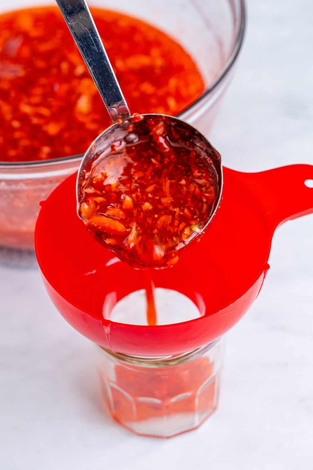 A ladle pouring in the strawberry jam through a funnel into a jar.