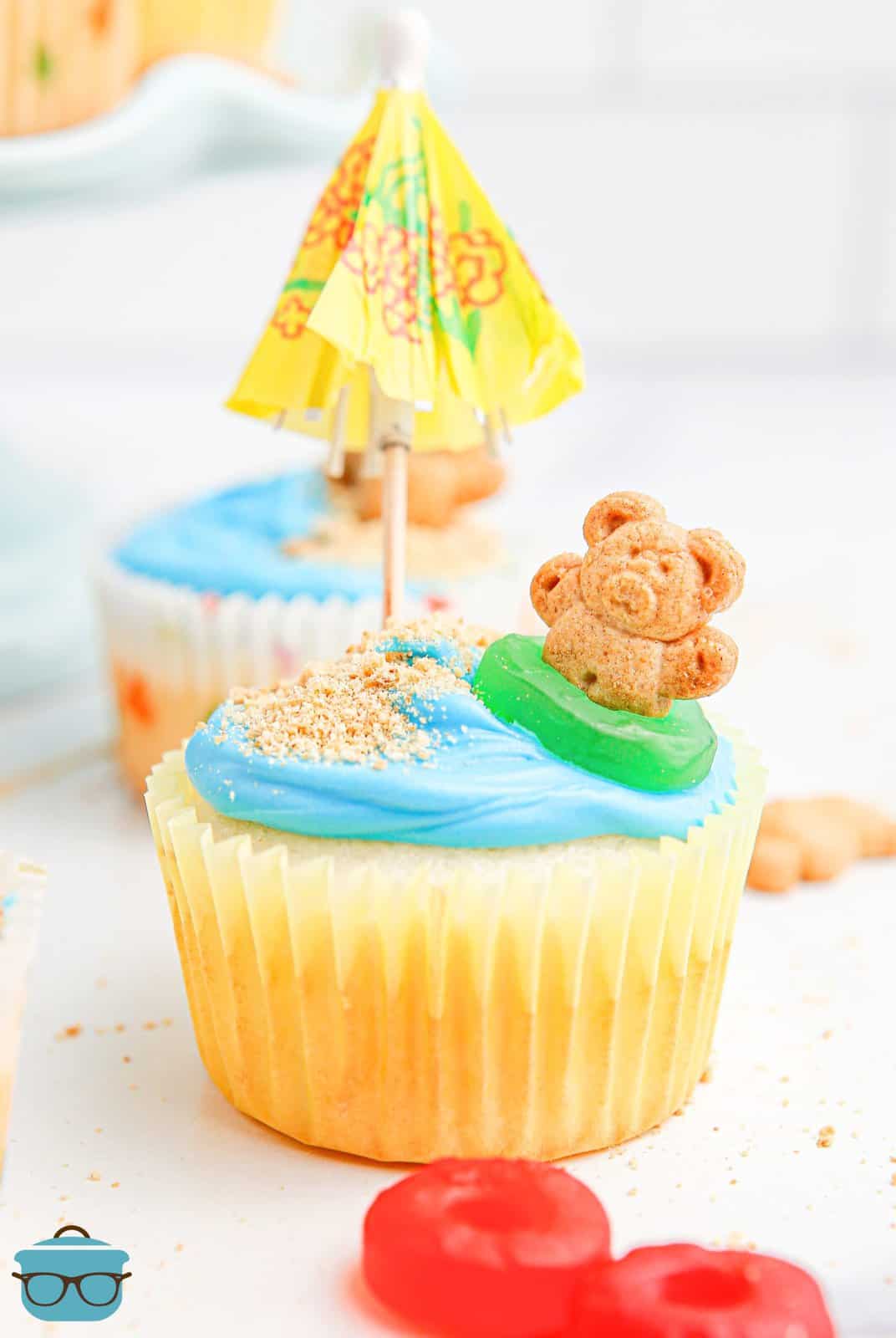 Looking closely at a cupcake decorated with a beach scene, umbrella, and teddy bear.