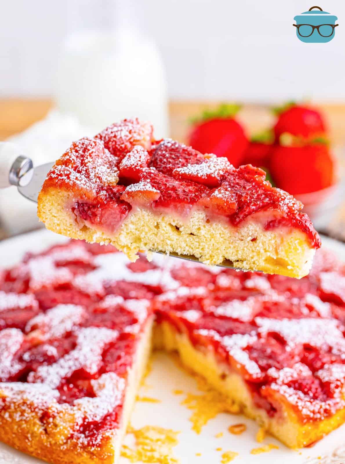 A slice of strawberry upside down cake on a serving utensil above the rest of the cake. 