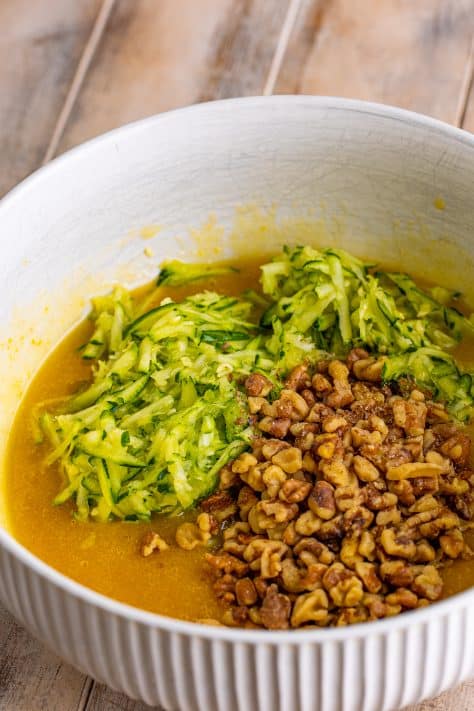 A mixing bowl with egg, egg yolk, oil, butter, brown sugar, granulated sugar, and vanilla, as well as walnuts and zucchini.
