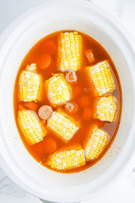 A slow cooker with water, Old Bay seasoning, hot sauce lemon juice, potatoes, bay leaves, sausage and corn cobs.