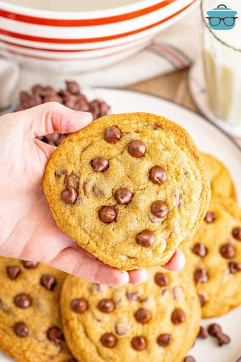 A hand holding a copycat Mrs. Fields Chocolate Chip Cookie above a plate of them.