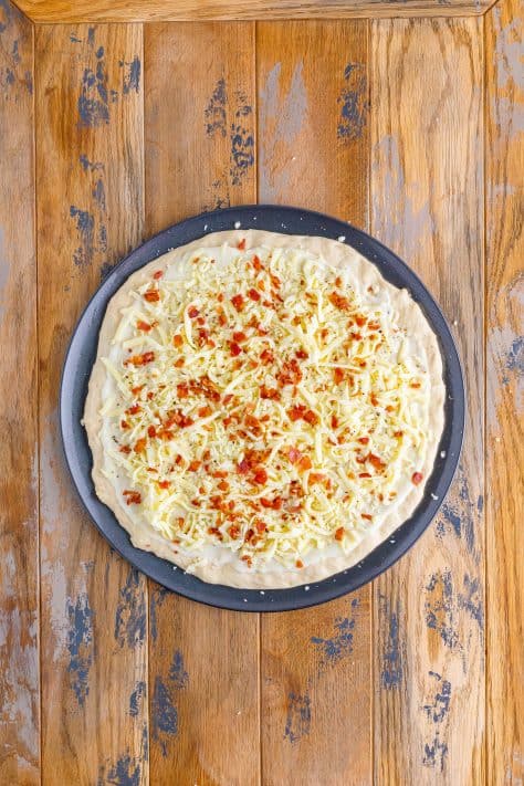 A pizza pan with dough, white sauce, cheese and bacon.