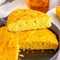 A slice of cornbread over a skillet filled with the rest of the loaf of cornbread.