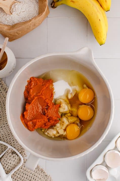 Bowl of a stand mixer with mashed banana, pumpkin puree, eggs, honey, sugar, and vegetable oil.