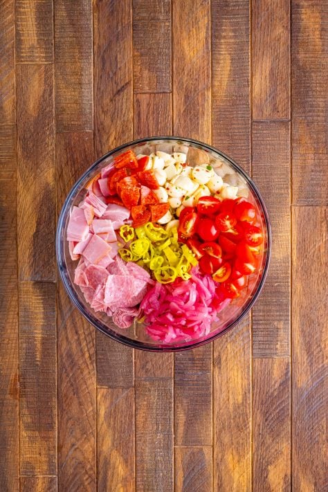 A large bowl of cooked rotini, cut up mozzarella balls, sliced pepperoncini peppers, cherry tomatoes, pickled red onion, salami, ham and pepperoni.