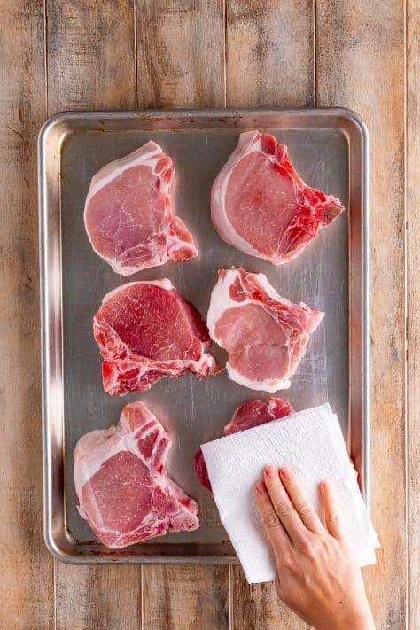 A baking sheet of pork chops with a hand patting them dry with a paper towel.