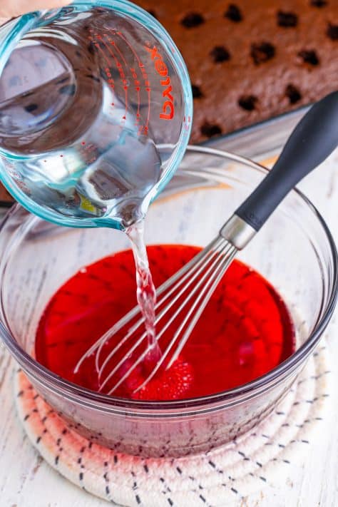 Cherry jello, a whisk, and water being poured in a mixing bowl.